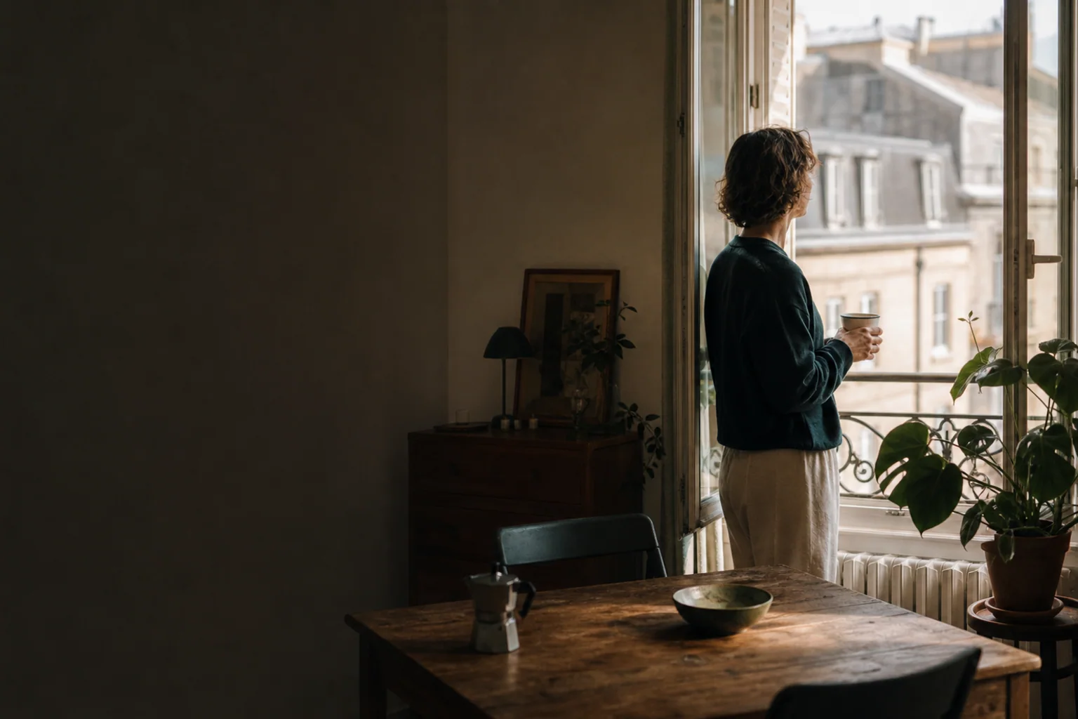 A person stands calmly by an open window holding a mug